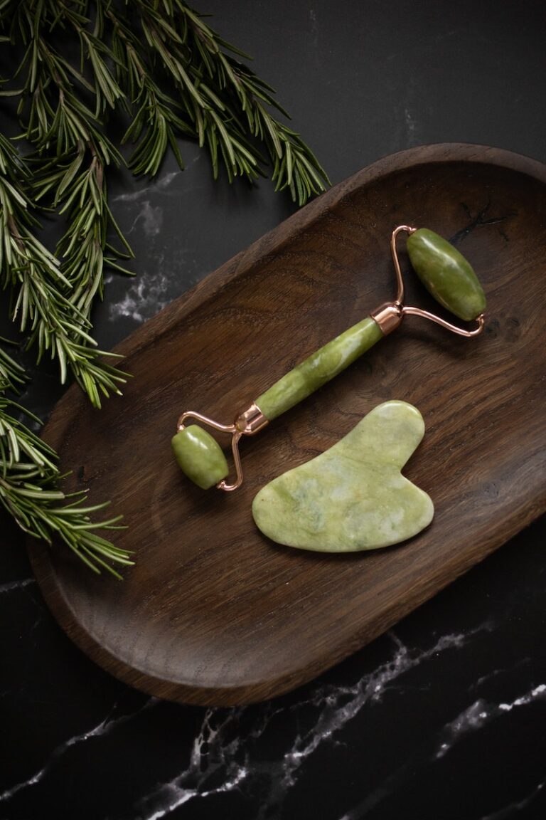 From above of Jade roller and Gua Sha massage tool placed on wooden plate on black marble table with fresh twigs of rosemary