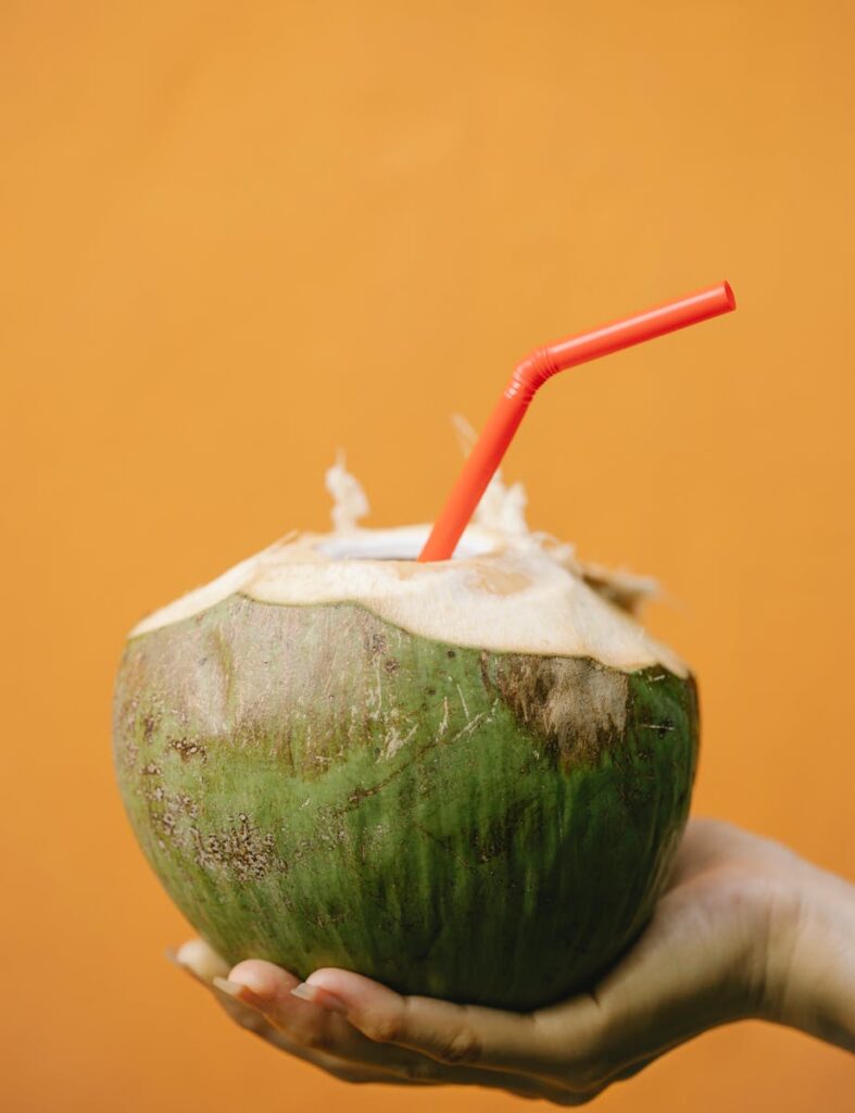 A hand holds a fresh coconut drink with a red straw against an orange background, exuding tropical vibes.