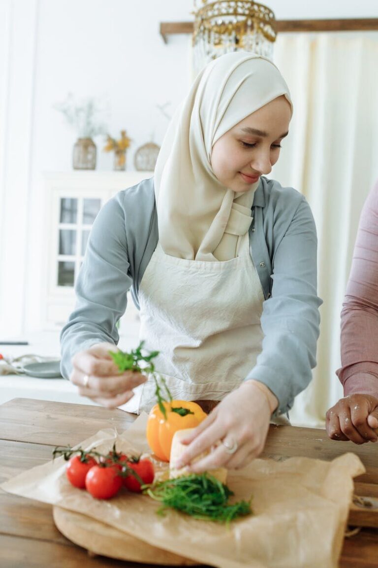 A woman wearing a hijab is preparing fresh vegetables at a wooden table indoors.