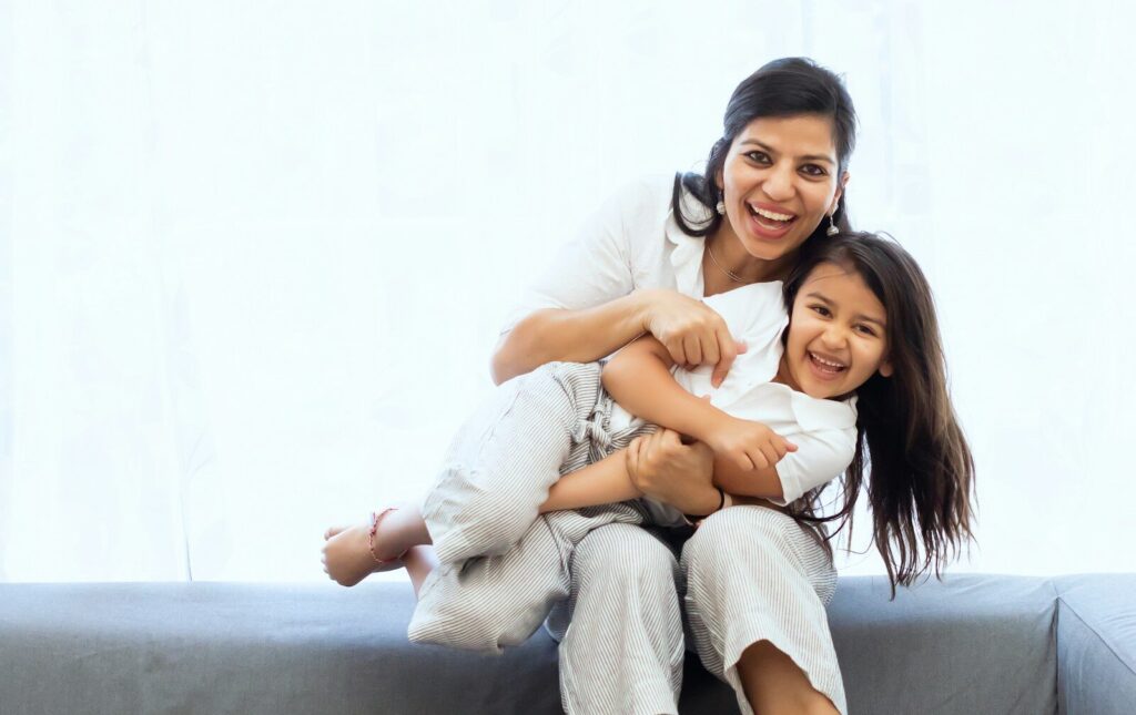 A woman holding a laughing child on a grey sofa in a bright room
