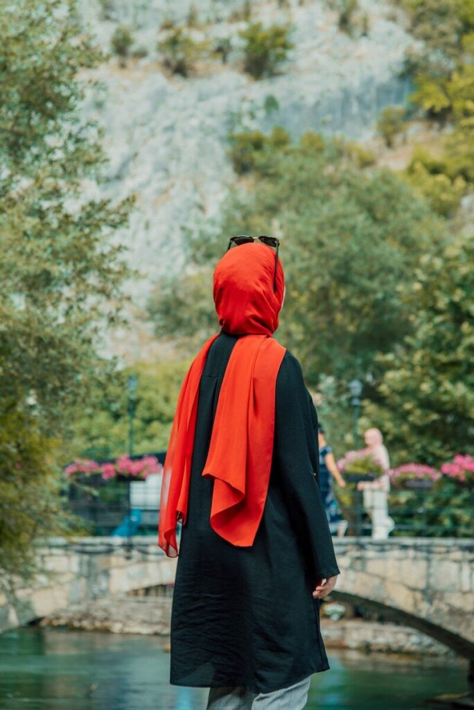 a person wearing a black robe and red head covering standing in front of a pond with trees and