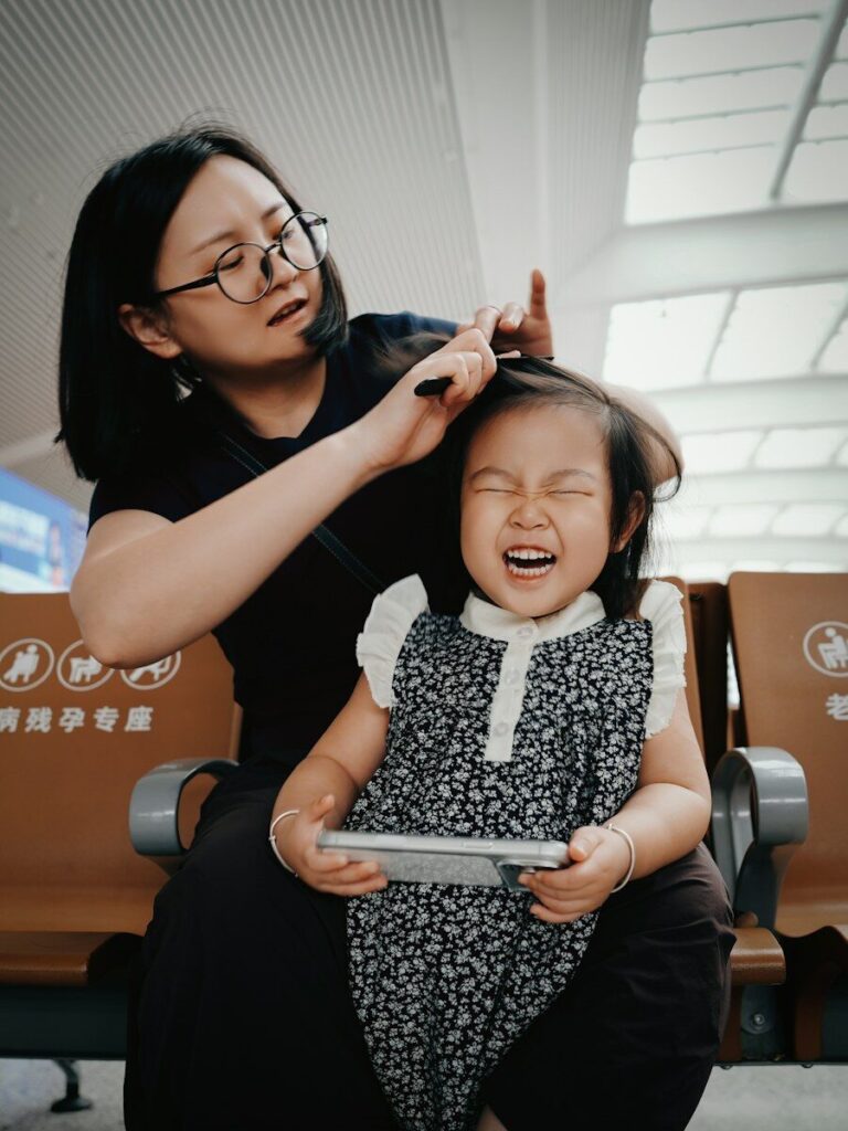 Mother combs daughter's hair, girl laughs holding smartphone