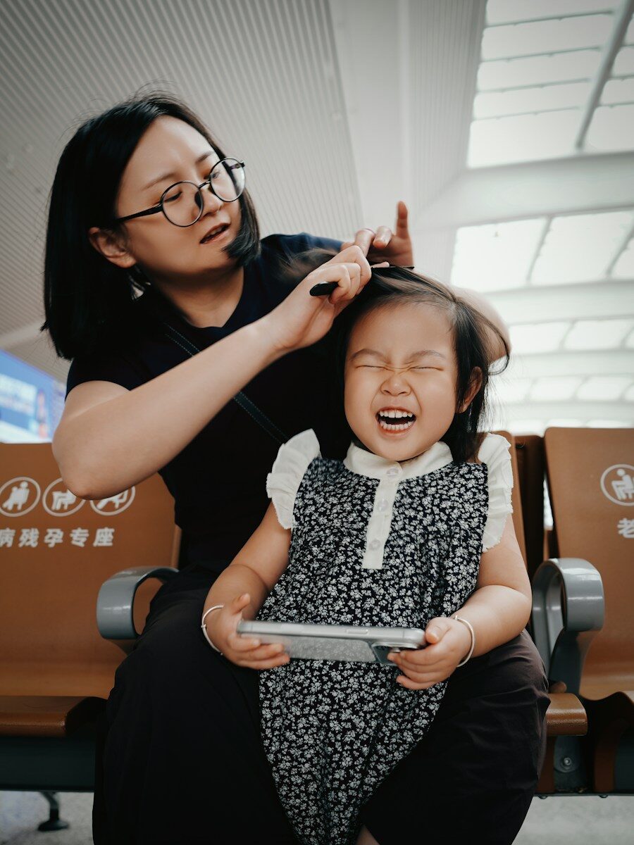 Mother combs daughter's hair, girl laughs holding smartphone